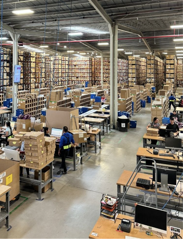 Workers pack boxes on tables in a large warehouse with shelves of products.
