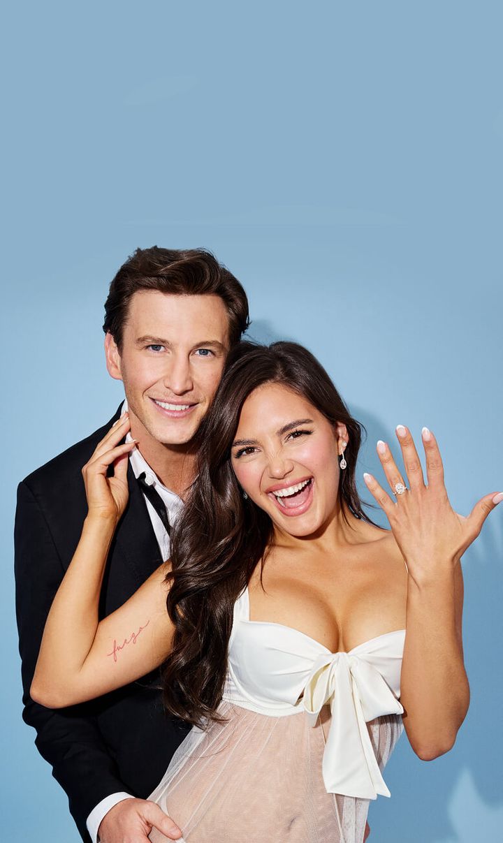 A man in a suit and a woman in a white dress show off an engagement ring.