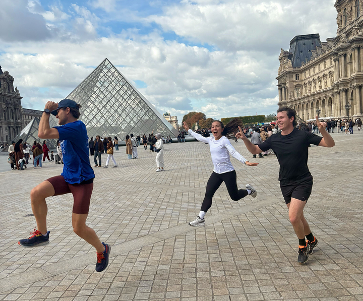 Three people leap joyfully in front of the Louvre Pyramid in Paris.