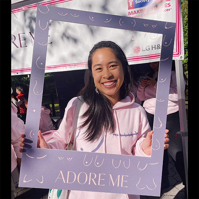 Woman in pink hoodie holds a "Adore Me" frame at a breast cancer event.