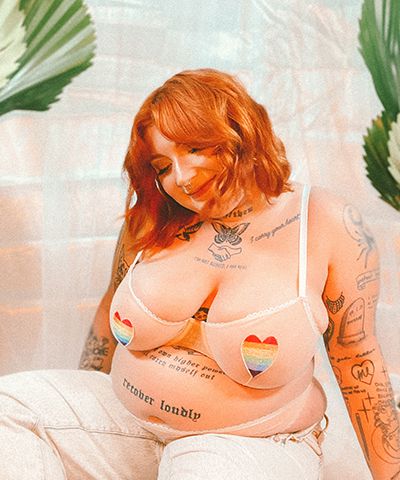 A person with tattoos and auburn hair wearing a white bra with rainbow hearts.
