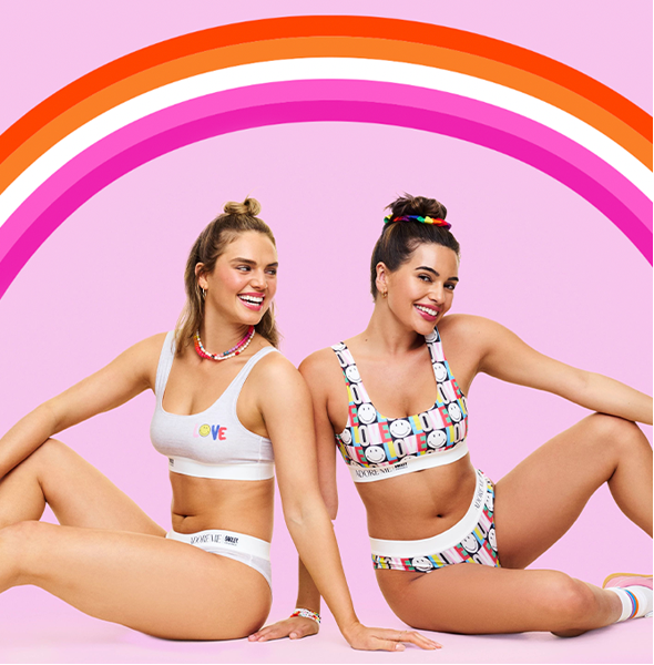 Two women model bralettes and underwear in front of an orange and pink rainbow.