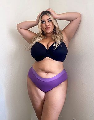 A curvy woman poses in a black bra and purple underwear.